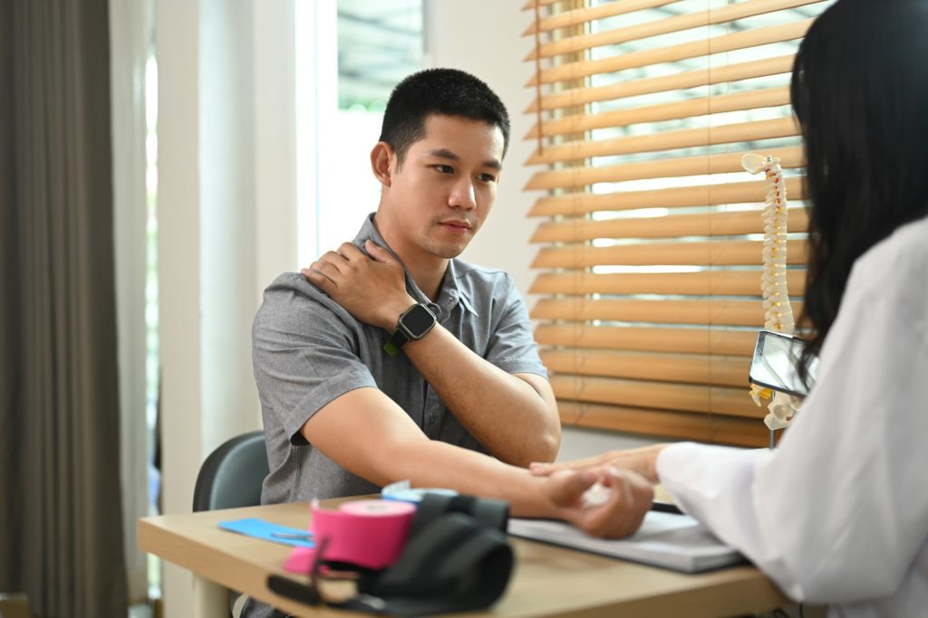 Male patient touching his shoulder describing symptoms to physiotherapist at rehabilitation clinic.